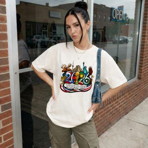May include: An off-white short-sleeve t-shirt featuring a colorful graphic of the 2026 World Cup with icons including a moose, a pyramid, the Statue of Liberty, and an eagle, with the text World Cup Canada Mexico USA printed below the design.