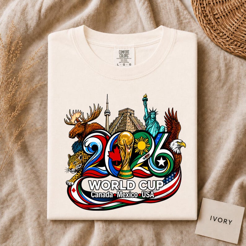 May include: An ivory t-shirt featuring a graphic print of the 2026 World Cup in Canada, Mexico, and the USA. The design includes the trophy, a moose, a jaguar, a bald eagle, the CN Tower, Chichen Itza, and the Statue of Liberty in vibrant colors.