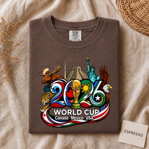 May include: A brown t-shirt featuring a graphic of the 2026 World Cup with the text Canada, Mexico, USA. The design includes a gold trophy, a moose, a jaguar, the Statue of Liberty, a bald eagle, and landmarks like the CN Tower and a pyramid.