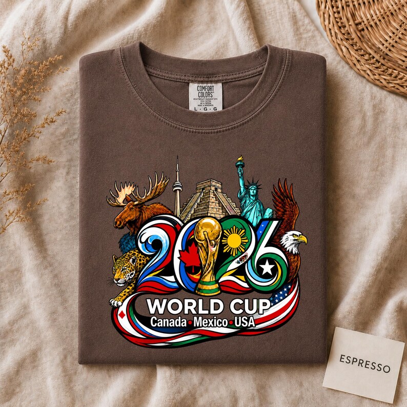 May include: A brown t-shirt featuring a graphic of the 2026 World Cup with the text Canada, Mexico, USA. The design includes a gold trophy, a moose, a jaguar, the Statue of Liberty, a bald eagle, and landmarks like the CN Tower and a pyramid.