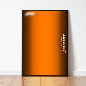 May include: A framed poster featuring an orange and brown gradient. The McLaren Formula 1 Team logo is displayed in white on the right side. The poster is in a black frame and is leaning against a white wall.
