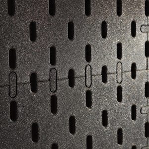 Eine schwarze Lochwand mit einem gleichmäßigen Raster aus vertikal ausgerichteten, abgerundeten rechteckigen Schlitzen. Die Oberfläche hat eine leicht texturierte, matte Optik und weist eine horizontale Nahtstelle zwischen zwei Paneelen auf.