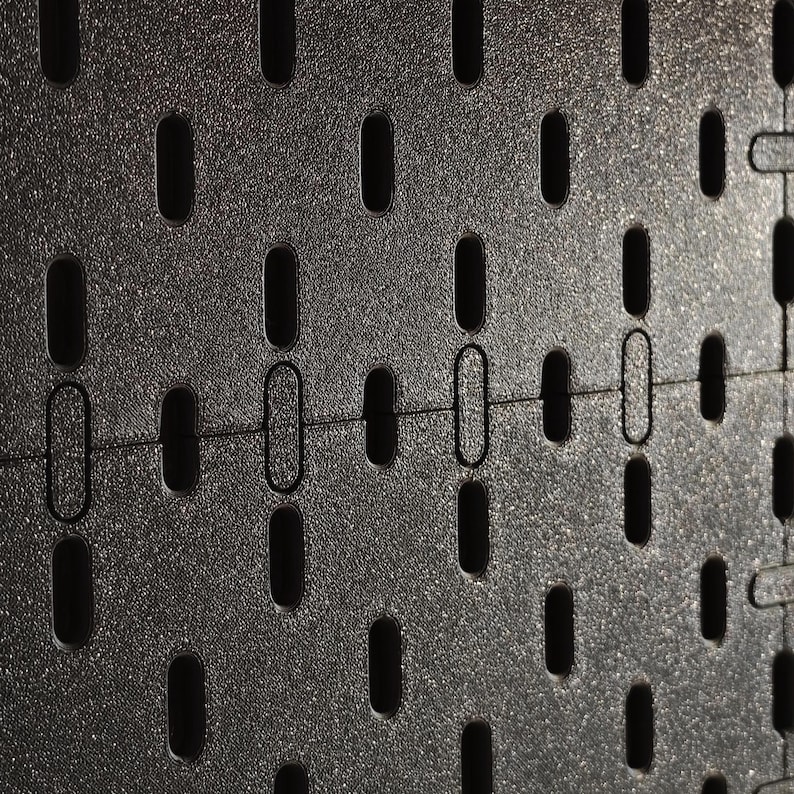 Eine schwarze Lochwand mit einem gleichmäßigen Raster aus vertikal ausgerichteten, abgerundeten rechteckigen Schlitzen. Die Oberfläche hat eine leicht texturierte, matte Optik und weist eine horizontale Nahtstelle zwischen zwei Paneelen auf.