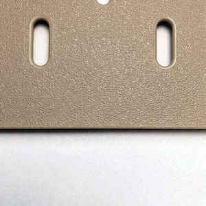 Eine beige rechteckige Platte mit einer texturierten, körnigen Oberfläche. Das Objekt verfügt über zwei vertikale, abgerundete rechteckige Schlitze, die symmetrisch angeordnet sind, sowie ein teilweises kreisförmiges Loch am oberen Rand.