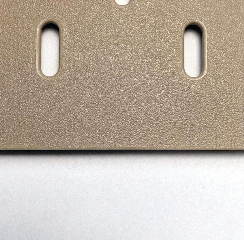 Eine beige rechteckige Platte mit einer texturierten, körnigen Oberfläche. Das Objekt verfügt über zwei vertikale, abgerundete rechteckige Schlitze, die symmetrisch angeordnet sind, sowie ein teilweises kreisförmiges Loch am oberen Rand.