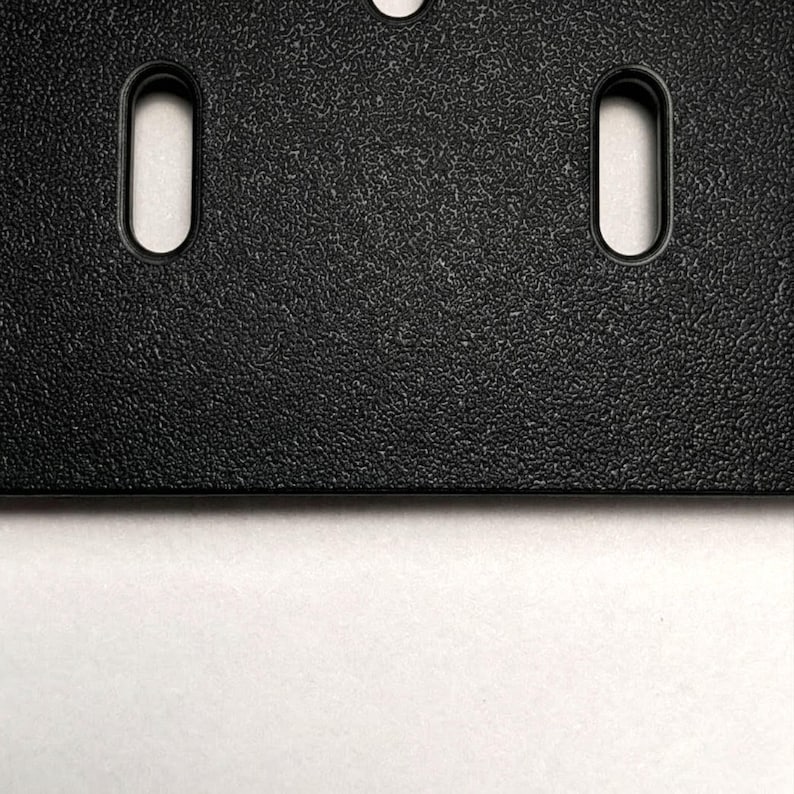 Eine schwarze Kunststoffplatte mit einer texturierten, matten Oberfläche, die zwei vertikale, abgerundete rechteckige Schlitze und ein einzelnes kreisförmiges Loch an der Oberkante aufweist. Die Platte hat eine gerade, saubere Unterkante.
