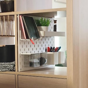 Eine weiße Lochwand zur Organisation in einem Holzregal. Sie verfügt über ein weißes Ablagefach mit einer grünen Topfpflanze und einem Glasgefäß, eine Metallhalterung für zwei Notizbücher sowie mehrere transparente und weiße Behälter mit Stiften.