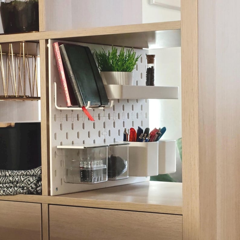 Eine weiße Lochwand zur Organisation in einem Holzregal. Sie verfügt über ein weißes Ablagefach mit einer grünen Topfpflanze und einem Glasgefäß, eine Metallhalterung für zwei Notizbücher sowie mehrere transparente und weiße Behälter mit Stiften.