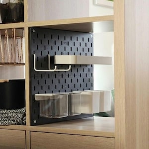Eine schwarze, rechteckige Lochplatte, die in einem Regal aus hellem Holz montiert ist. Die Lochplatte verfügt über ein Raster aus vertikalen Schlitzen und ist mit weißen Metallhaken, einem weißen rechteckigen Regal und zwei durchsichtigen Kunststoffbehältern ausgestattet.