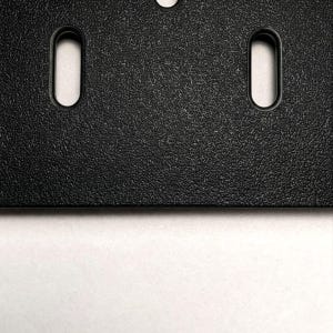 Eine schwarze Kunststoffplatte mit texturierter, matter Oberfläche, die vertikale, längliche ovale Befestigungsschlitze aufweist. Die Platte hat eine gerade Unterkante und eine gleichmäßige, körnige Oberflächenstruktur auf dem gesamten Bauteil.