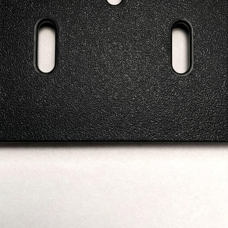 Eine schwarze Kunststoffplatte mit texturierter, matter Oberfläche, die vertikale, längliche ovale Befestigungsschlitze aufweist. Die Platte hat eine gerade Unterkante und eine gleichmäßige, körnige Oberflächenstruktur auf dem gesamten Bauteil.