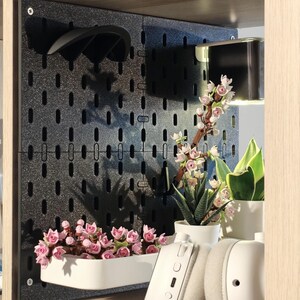 Vorschau: Eine schwarze Lochplatte mit glitzerndem Finish, montiert in einem Holzregal. Sie verfügt über eine weiße rechteckige Ablage mit rosafarbenen blütenförmigen Bausteinen, einen schwarzen gebogenen Haken und eine kleine Leuchte.