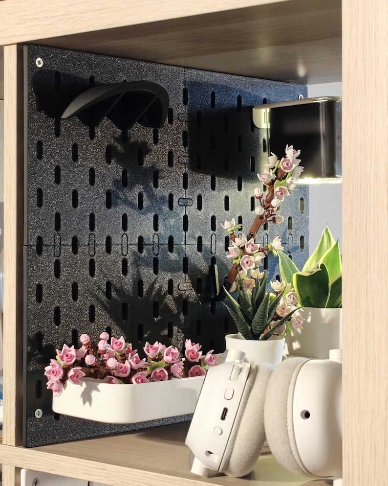 Vorschau: Eine schwarze Lochplatte mit glitzerndem Finish, montiert in einem Holzregal. Sie verfügt über eine weiße rechteckige Ablage mit rosafarbenen blütenförmigen Bausteinen, einen schwarzen gebogenen Haken und eine kleine Leuchte.