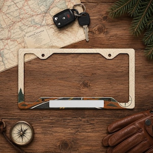 May include: A rectangular license plate frame featuring a topographic map pattern in beige and brown, with a dark green pine tree graphic on the left side and a compass rose detail on the right, designed for standard vehicle license plates.