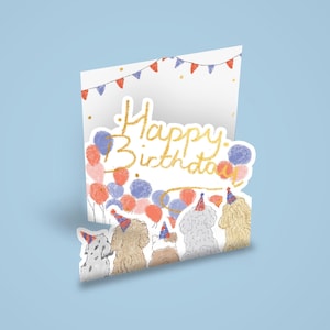 Op de afbeelding: Een pop-up verjaardagskaart met een lichtblauwe achtergrond. De kaart toont de woorden "Happy Birthday" in goudglitter, ballonnen en illustraties van honden met feesthoeden. Rode, blauwe en roze ballonnen zijn verspreid.