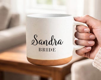 May include: A ceramic mug with a speckled off-white upper body and a solid terracotta-colored base. The front features the name Sandra in a black cursive script font, with the word BRIDE printed in a black serif font directly underneath it.