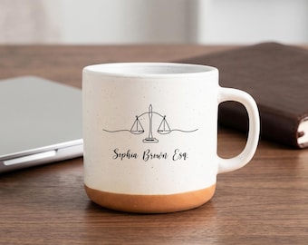 Può includere: Una tazza in ceramica bianca con finitura maculata e base color terracotta. La parte anteriore presenta un'illustrazione a linee nere di una bilancia della giustizia e il testo stampato "Sophia Brown Esq." in carattere corsivo.
