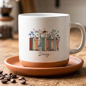 Könnte beinhalten: Ein gesprenkelter weißer Keramikbecher mit Henkel, verziert mit einer bunten Illustration einer Bücherreihe mit Wildblumen und dem in schwarzer Schreibschrift gedruckten Namen Daisy. Der Becher steht auf einer passenden Untertasse aus Terrakotta.