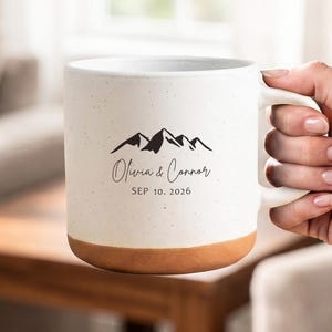 Puede incluir: Una taza de cerámica color crema con acabado moteado y base color terracota. El frente presenta un gráfico de montaña negro sobre el texto "Olivia & Connor" y la fecha "SEP 10. 2026" impresos en una fuente oscura y clara.