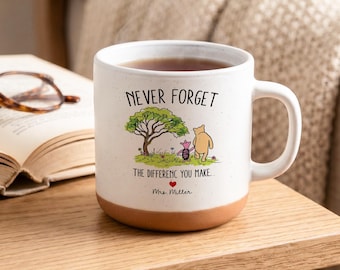 May include: A white ceramic mug with a speckled finish and a terracotta-coloured base. The mug features a printed illustration of Winnie the Pooh and Piglet walking under a tree, with the text NEVER FORGET THE DIFFERENC YOU MAKE and Mrs. Miller.