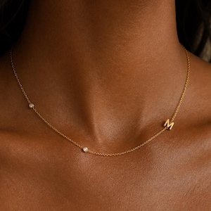 Op de afbeelding: Een delicate gouden ketting met een kleine initiaal "M" hanger en drie kleine, sprankelende edelstenen. De ketting wordt getoond op de hals van een persoon, waarbij het elegante ontwerp en de subtiele glans worden benadrukt. De ketting is dun en de hanger is klein.