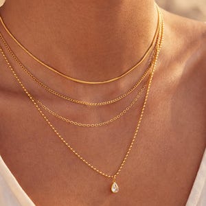 May include: Four gold necklaces of varying styles. The top necklace is a fine chain, followed by a curb chain, a delicate chain, and a beaded chain with a teardrop pendant. The necklaces are layered and worn together.