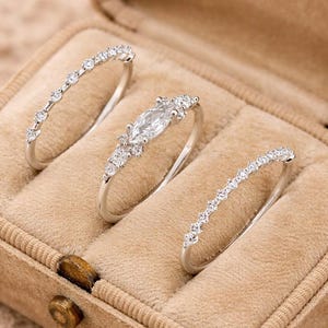 Könnte beinhalten: Drei silberne Ringe mit klaren Edelsteinen, präsentiert in einer beigen Schmuckschatulle. Die Ringe weisen verschiedene Designs auf, darunter ein Stein im Marquise-Schliff und zarte Bänder mit kleinen, runden Edelsteinen. Die Schatulle hat ein weiches, samtartiges Innenfutter.