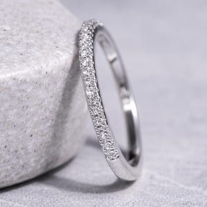 Op de afbeelding: Een zilveren ring met een band ingelegd met kleine, heldere edelstenen. De ring leunt tegen een grijs, getextureerd oppervlak. De edelstenen schitteren en reflecteren licht. Het is een sieraad.