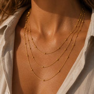 Könnte beinhalten: Eine mehrreihige Goldhalskette bestehend aus drei zarten Ketten in unterschiedlichen Längen. Jede Kette ist mit kleinen, gleichmäßig verteilten Goldperlen besetzt, die einen geschichteten Effekt über dem Schlüsselbein und dem Brustbereich erzeugen.