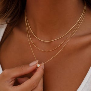 Può includere: Un set di tre collane d'oro. La collana superiore è una catena a serpente, quella centrale è una catena di perline e quella inferiore presenta un piccolo pendente rotondo con una pietra trasparente. Le collane sono indossate sul collo.