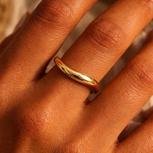 May include: A gold ring with a smooth, curved design. The ring is worn on a finger, highlighting its polished surface and elegant shape. The ring's design is simple yet eye-catching, reflecting light and adding a touch of sophistication.