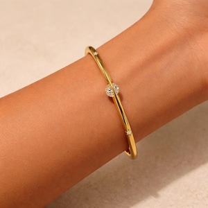 Könnte beinhalten: Ein goldfarbenes Armreif-Armband mit einem zentralen Cluster aus klaren Kristallen und kleineren Kristallen entlang des Bandes. Das Armband wird an einem Handgelenk getragen und zeigt sein elegantes Design und seinen Glanz. Ein schlichtes und stilvolles Schmuckstück.