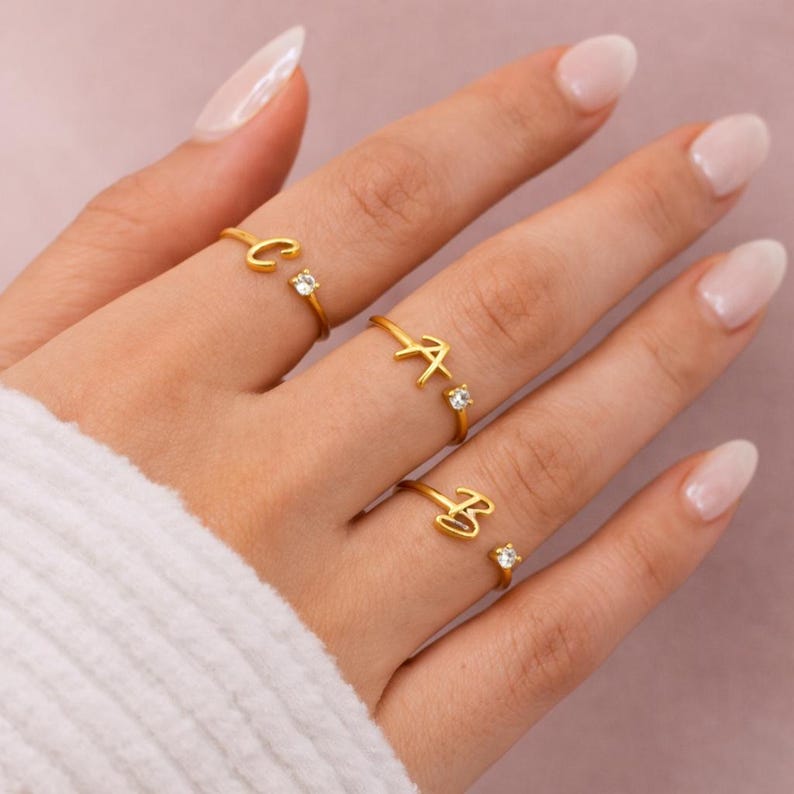 May include: Gold-coloured initial rings with small, clear gemstones. The rings feature the letters C, A, and B. The rings are on a hand with manicured nails, and the background is a soft pink.