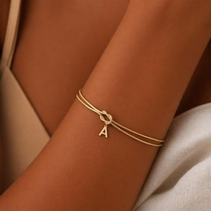 Può includere: Un braccialetto dorato con un design a nodo e un ciondolo con la lettera "A". Il braccialetto presenta un design a doppio filo e viene indossato sul braccio. Un gioiello delicato.