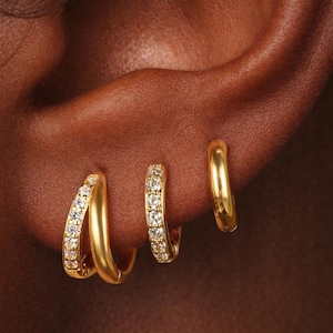 May include: A close-up shot of three gold hoop earrings. Two are adorned with small, clear gemstones, and the third is a plain gold hoop. The earrings are shown on an ear, highlighting their design and the way they sit.