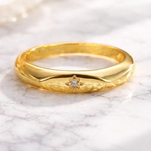 Könnte beinhalten: Ein goldfarbener Ring mit einem Sternmuster und einem kleinen, klaren Edelstein in der Mitte. Der Ring hat eine glatte, polierte Oberfläche und ein leicht abgerundetes Band. Der Ring wird auf einer weiß-grauen Marmoroberfläche präsentiert.