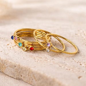 Può includere: Una collezione di anelli color oro con pietre preziose colorate. Gli anelli sono impilati, alcuni con una singola gemma, altri semplici. Le pietre preziose sono blu, rosa, verdi, gialle, rosse, viola e trasparenti.