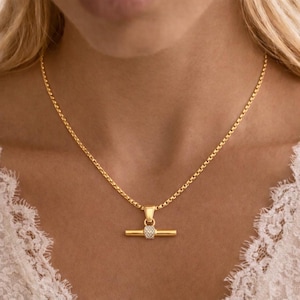 May include: A gold necklace with a bar pendant. The pendant has a small, round cluster of clear stones in the centre of the bar. The chain is a delicate box chain style.