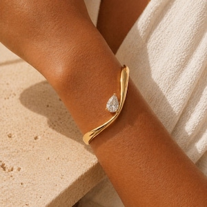 Könnte beinhalten: Ein goldfarbenes Armreif-Armband mit einem birnenförmigen, klaren Edelstein. Das Armband schmiegt sich um das Handgelenk, wobei der Edelstein am äußeren Rand angebracht ist. Der Schmuck wird am Arm einer Person gezeigt.