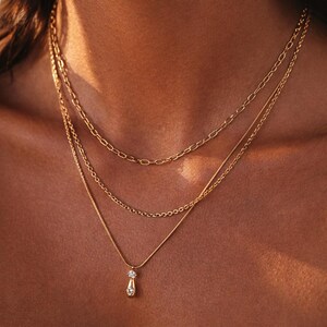 Op de afbeelding: Een gelaagde set van drie gouden kettingen. De bovenste ketting heeft een schakelontwerp, de middelste is een delicate ketting en de onderste ketting heeft een hanger met twee heldere edelstenen. De kettingen worden op de huid van een persoon getoond.