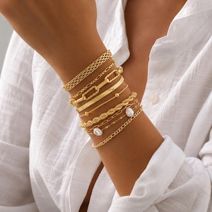 May include: A set of gold-toned bracelets worn together, featuring various chain styles including a flat herringbone chain, a twisted rope chain, a link chain with rectangular segments, and two delicate chains accented with small white pearls.