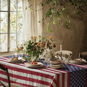 Op de afbeelding: Een rechthoekig tafelkleed met het patroon van de Amerikaanse vlag met rode en witte strepen en een blauw veld met witte sterren. De tafel is gedekt met witte keramische borden, heldere glazen en een groot bloemstuk met oranje bloemen.