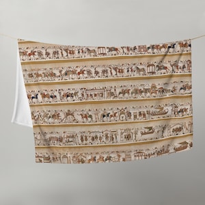 May include: A beige throw blanket featuring a detailed printed reproduction of the Bayeux Tapestry. The design displays multiple horizontal bands filled with historical scenes of figures on horseback, ships, and architectural structures in muted tones.