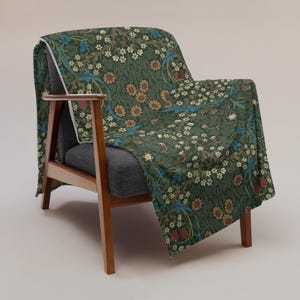 May include: A dark green throw blanket featuring a dense botanical pattern of small white flowers, orange blossoms, and blue leaves draped over a dark gray armchair with wooden armrests and legs. The fabric has a white edge trim along the side.
