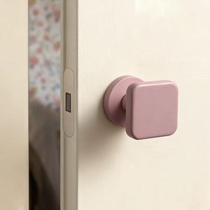 Può includere: Una maniglia quadrata rosa con base circolare è montata su una porta color crema. La porta è leggermente socchiusa, rivelando uno scorcio di una carta da parati a motivi floreali sullo sfondo.