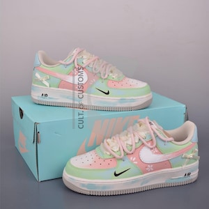 Puede incluir: Zapatillas personalizadas en colores pastel con detalles florales y el logo de Nike. Las zapatillas tienen una base blanca con tonos rosas, verdes y azules, y el texto "AID" es visible en el lateral. Los cordones son de color rosa claro.