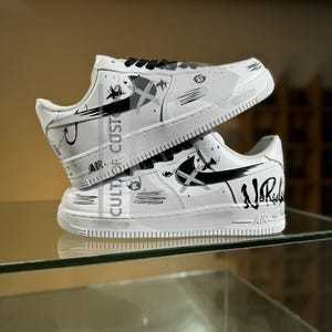 May include: A pair of white trainers with black laces and black graphic designs featuring abstract shapes, stars, and the text "No Rules" on the side. The shoes have a white sole, a black swoosh logo, and the word "AIR" embossed on the heel area.