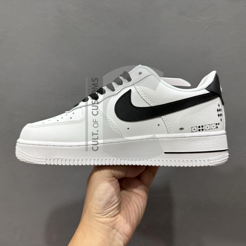 Peut inclure: Une basket Nike Air Force 1 blanche et noire. La chaussure pr&eacute;sente une tige en cuir blanc avec un logo Nike noir sur le c&ocirc;t&eacute;. La semelle est blanche et les lacets sont gris. La chaussure a un design bas et un style athl&eacute;tique classique.