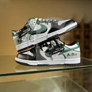 Op de afbeelding: Een paar op maat gemaakte sneakers, op elkaar gestapeld. De schoenen zijn wit, zwart en grijs met groene details. De Nike swoosh is groen. De tekst "Be Strong" is zichtbaar op één schoen.