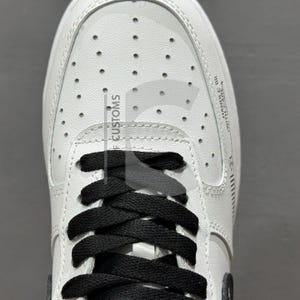 Peut inclure: Gros plan d'une basket en cuir blanc avec des lacets noirs. La chaussure porte l'inscription "F CUSTOMS" et "HANDLE WITH CARE" sur le c&ocirc;t&eacute;. Le design comprend de petites perforations sur le dessus.